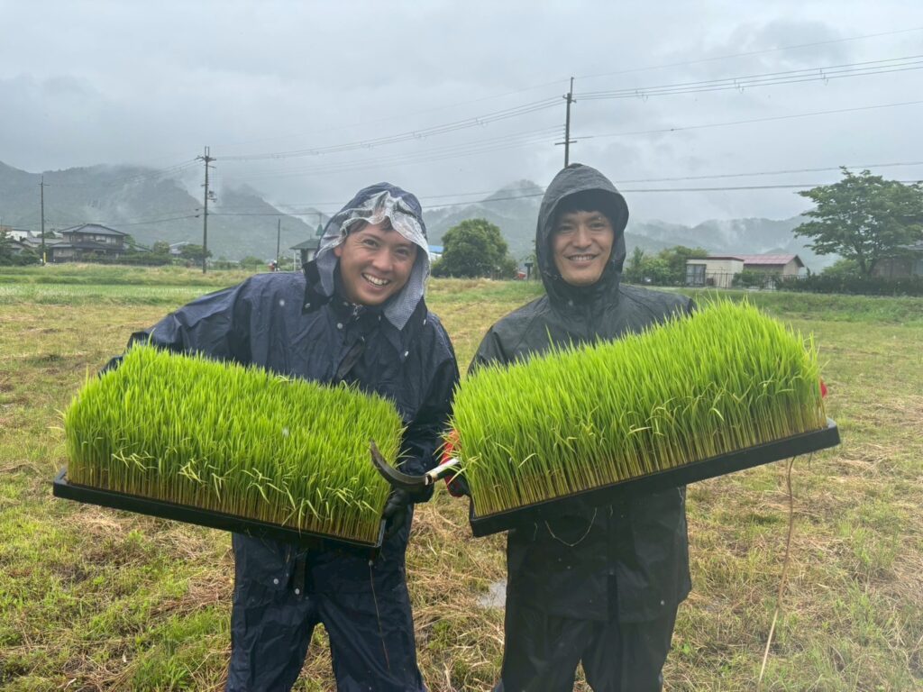 雨の中、稲の苗を手に笑顔を見せる参加者（firva農業プロジェクト）